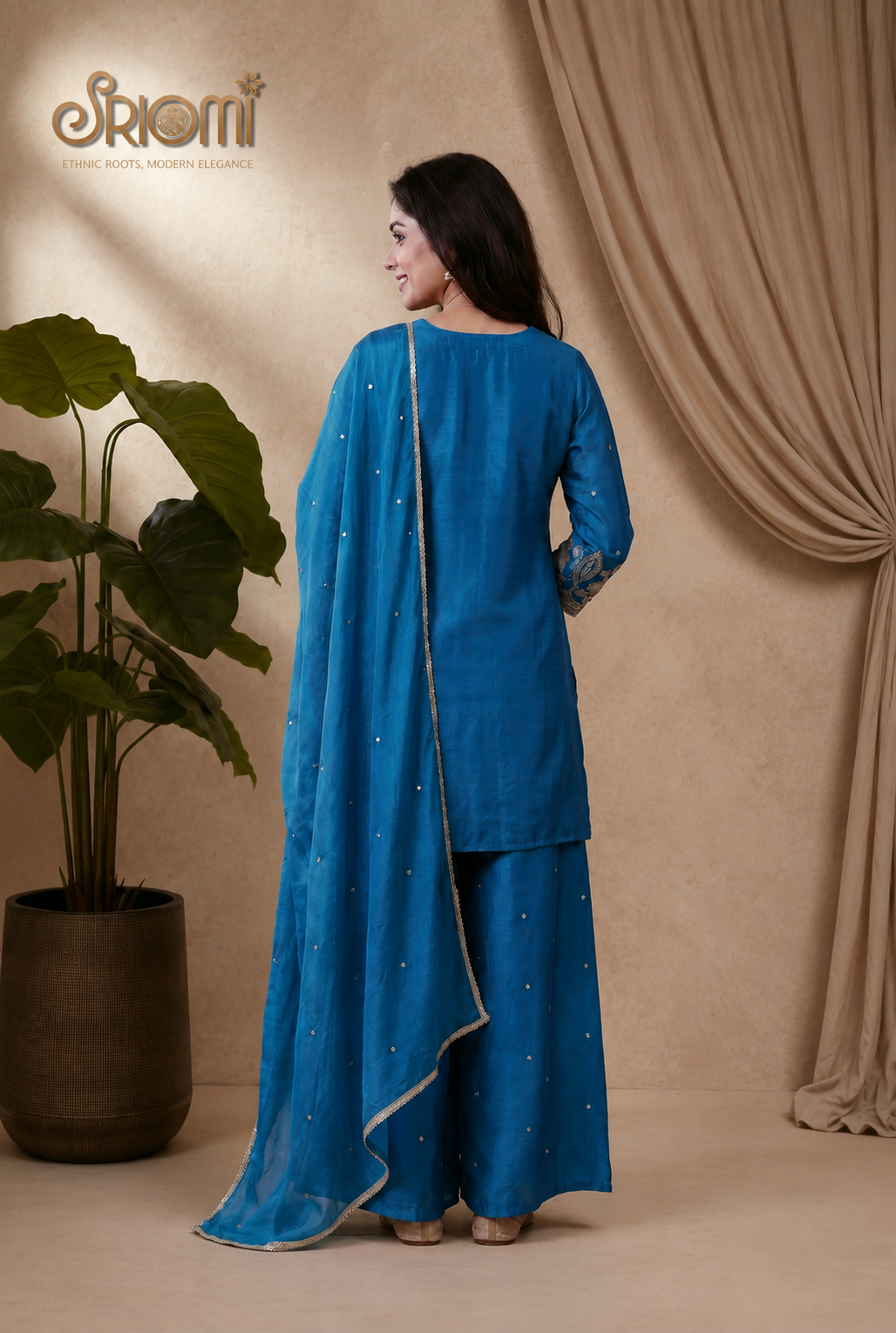 Woman in a blue traditional outfit standing against a beige curtain with 'Srioni' branding.