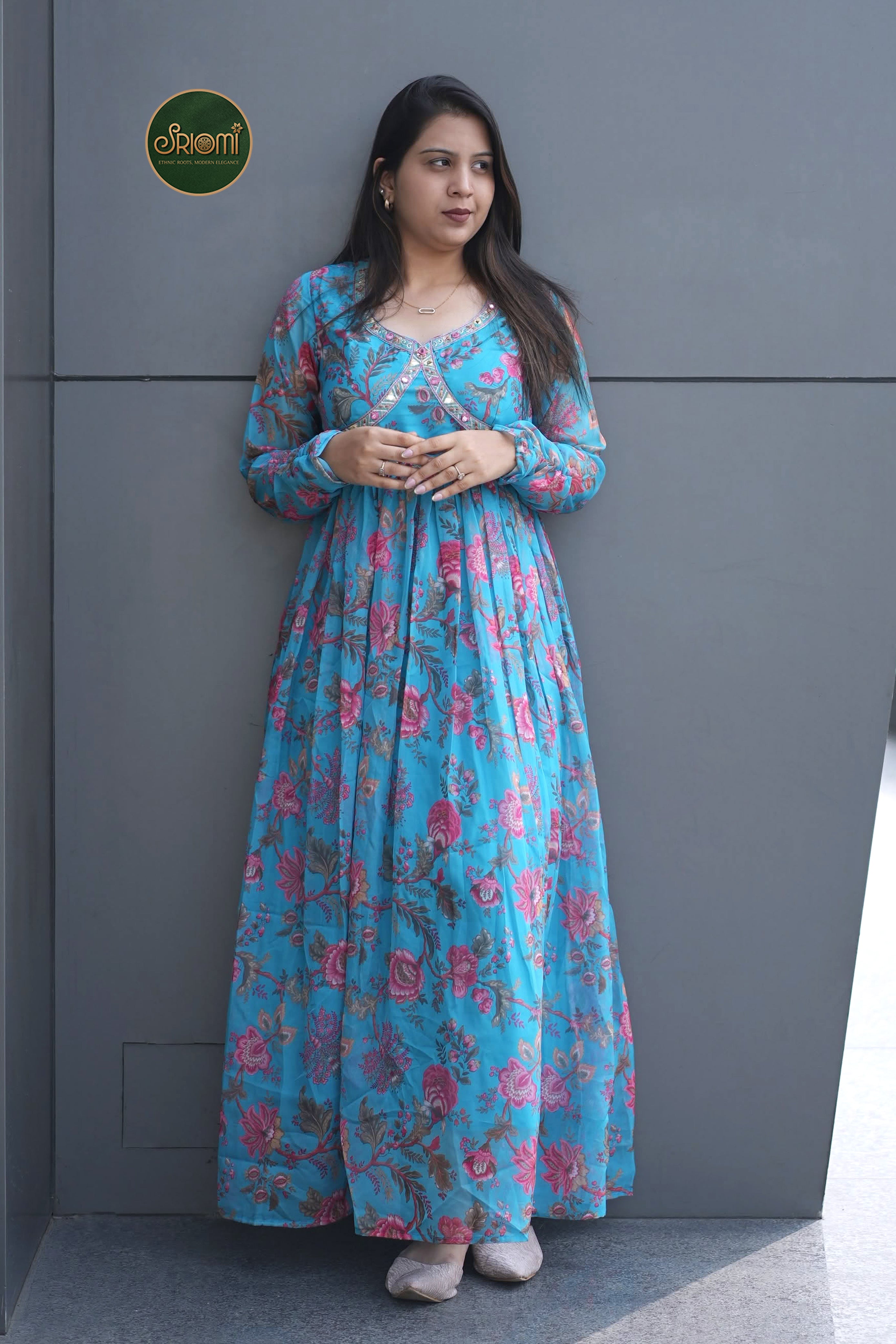 A woman wearing a long-sleeved, floor-length blue maxi dress with a pink floral pattern, standing against a dark gray paneled wall.