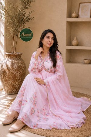 A woman wearing a light pink floral printed Anarkali gown sits on a woven mat in a warm, earthy-toned room. The dress features a V-neckline, long sleeves, and a flowing dupatta.