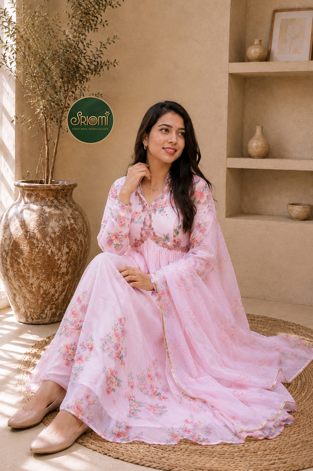 A woman wearing a light pink floral printed Anarkali gown sits on a woven mat in a warm, earthy-toned room. The dress features a V-neckline, long sleeves, and a flowing dupatta.
