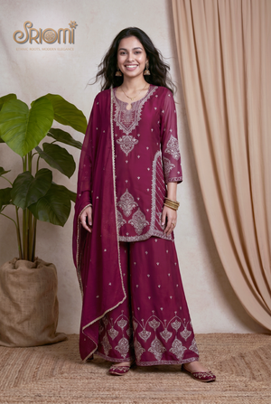 Woman in a maroon traditional outfit with intricate patterns, standing against a beige curtain.