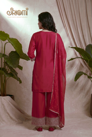 Person wearing a red traditional outfit with a dupatta, standing against a neutral background with plants.