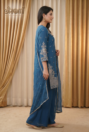 Woman wearing a blue embroidered outfit standing in front of beige curtains