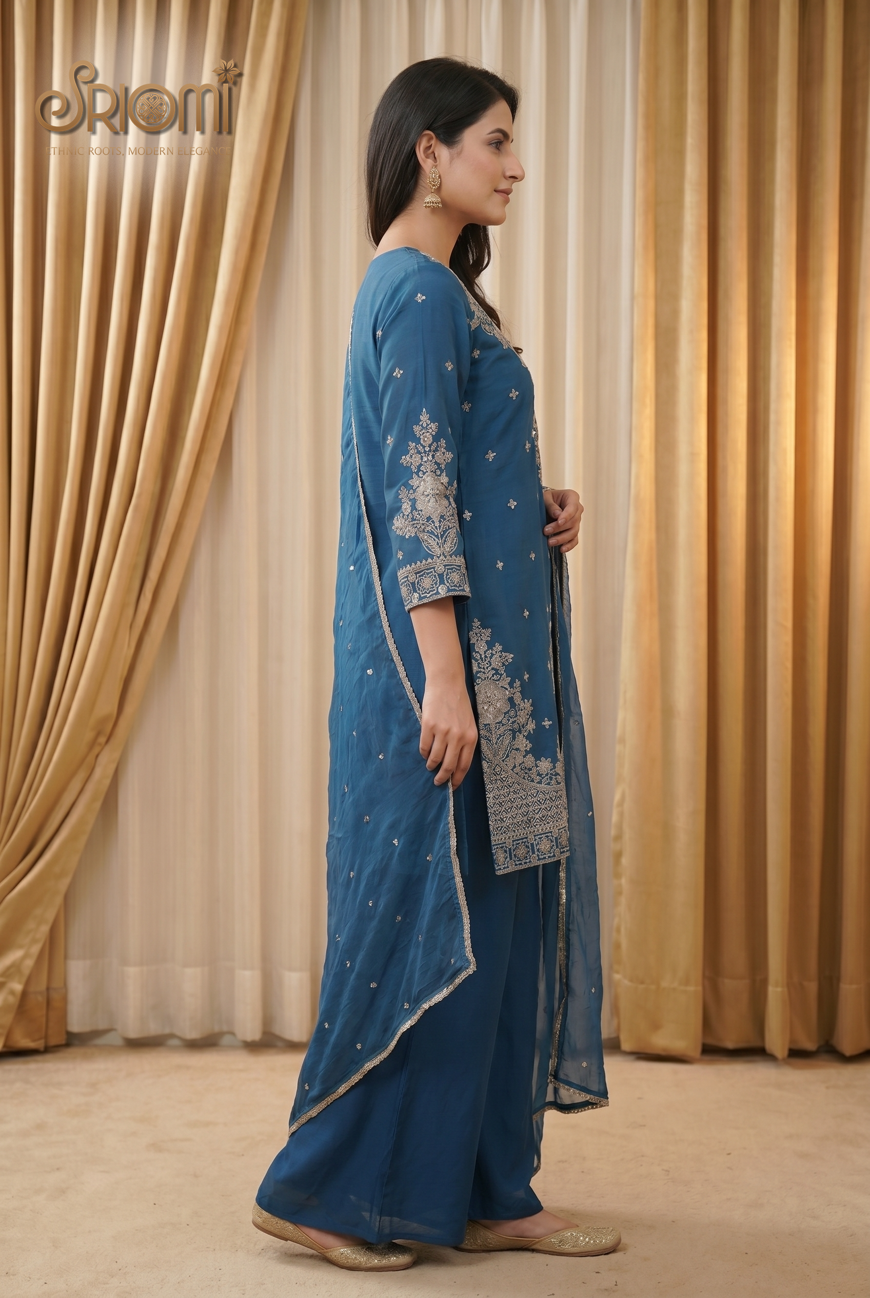 Woman wearing a blue embroidered outfit standing in front of beige curtains