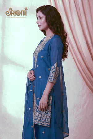 Woman wearing a blue traditional outfit with gold embroidery against a light pink curtain.
