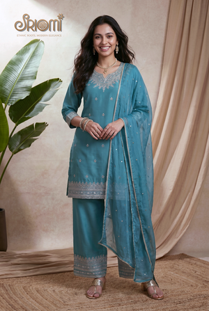 Woman wearing a teal traditional outfit with silver details, standing against a beige curtain.