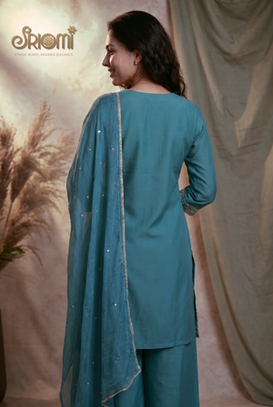 Woman wearing a teal traditional outfit with a matching dupatta against a beige curtain background.