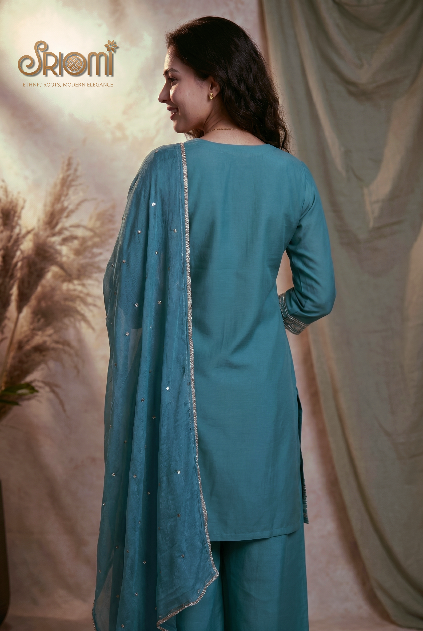 Woman wearing a teal traditional outfit with a matching dupatta against a beige curtain background.
