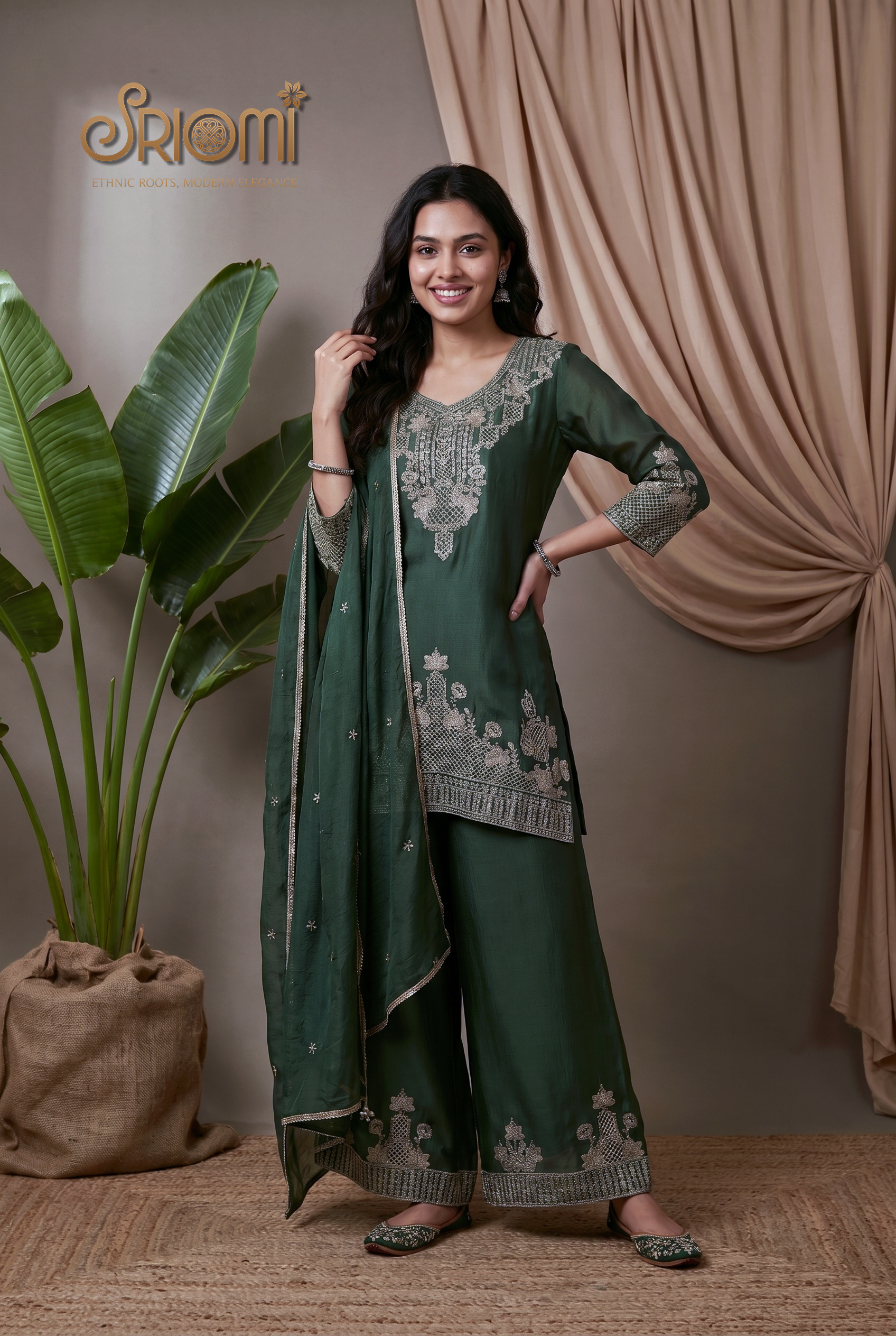 Woman wearing a green traditional outfit with embroidery, standing in front of beige curtains and a plant.